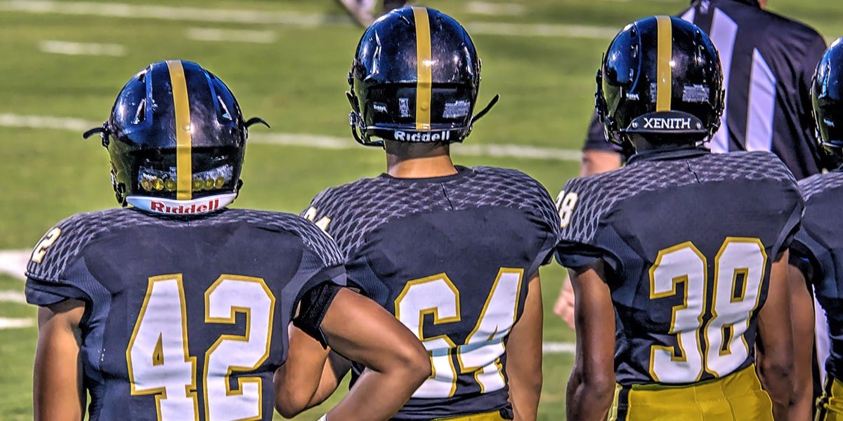3 high school football players in uniform with helmets on, standing on sideline of football field.